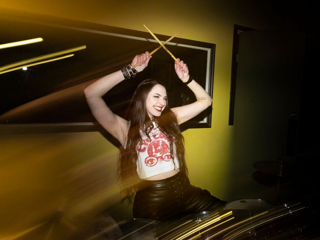 Energetic female drummer raises drumsticks overhead under golden stage lighting in dynamic music photo.