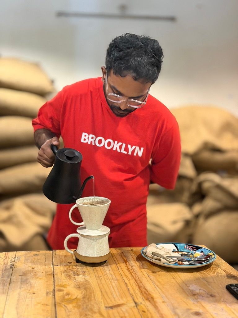 Man in red Brooklyn t-shirt pouring hot water into a pour-over coffee filter on a wooden table.