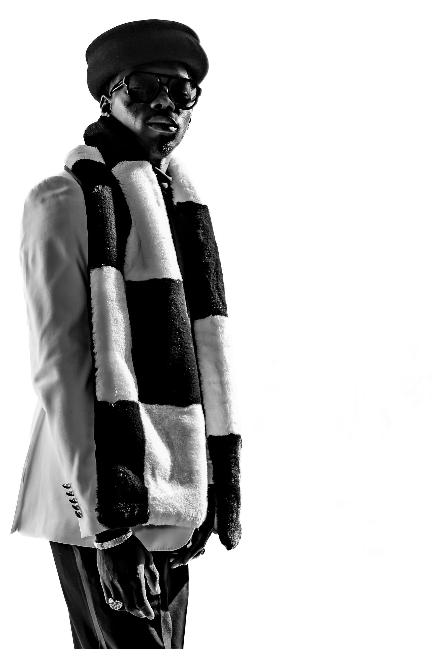 Black and white portrait of a man wearing a striped scarf, blazer, cap, and sunglasses against white background.