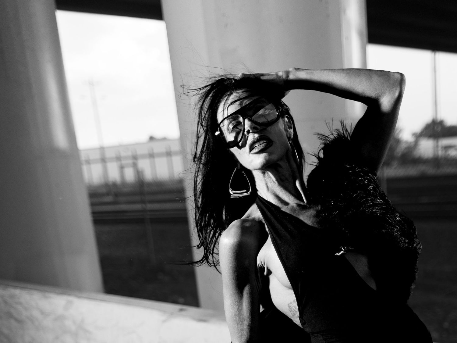 Black and white fashion portrait of woman in black dress posing under urban overpass structure.