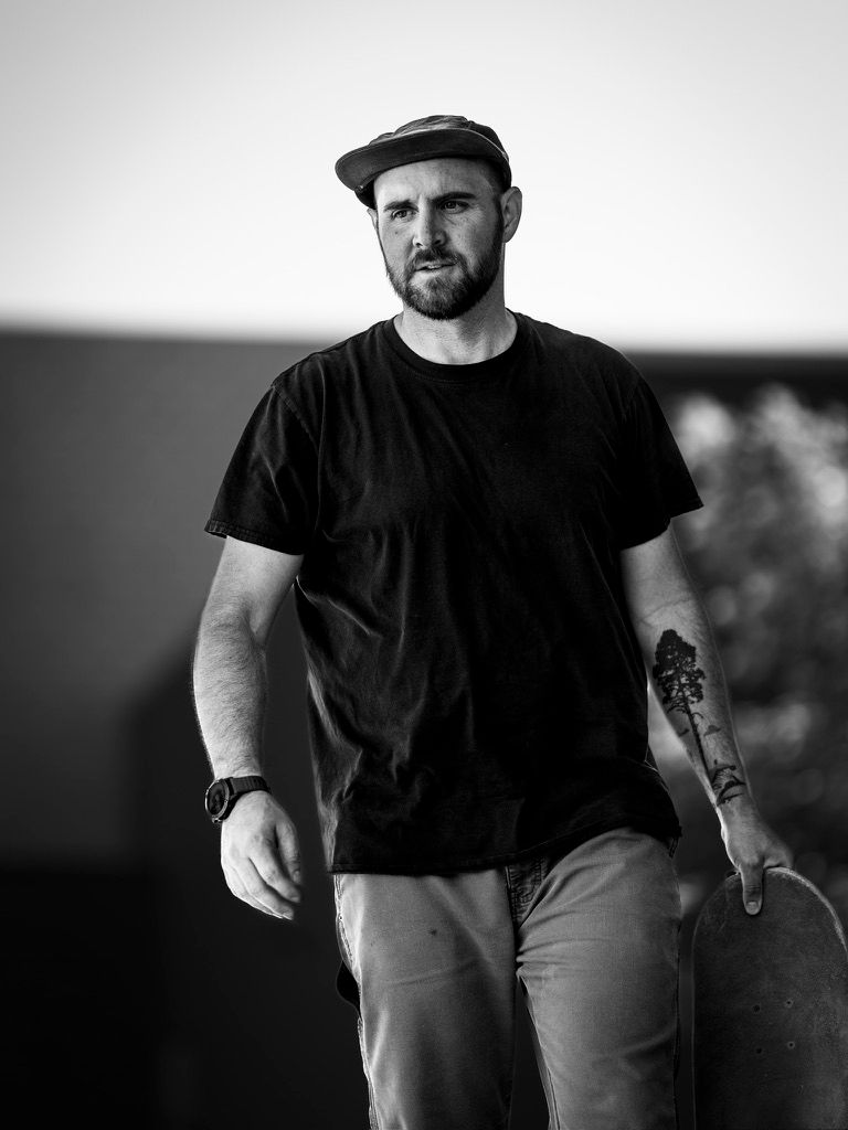 Bearded man in black t-shirt and flat cap holds skateboard outdoors in black and white portrait photo.