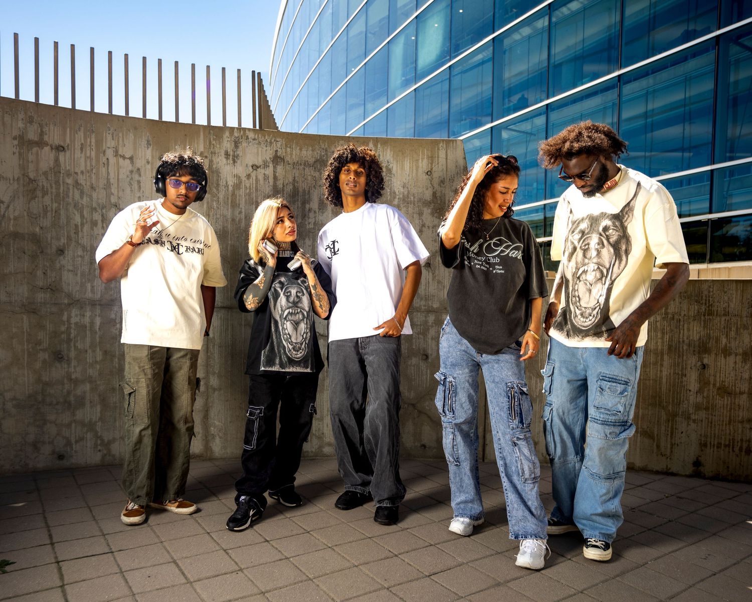 Five stylish young adults pose outdoors near a modern glass building wearing urban streetwear fashion.