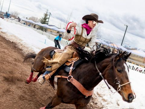Female cowgirl riding dark horse at winter rodeo competition in snowy arena.
