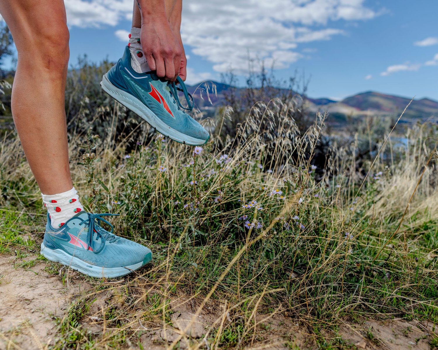 Person tying teal running shoes on a trail with green grass and mountain landscape in the background.
