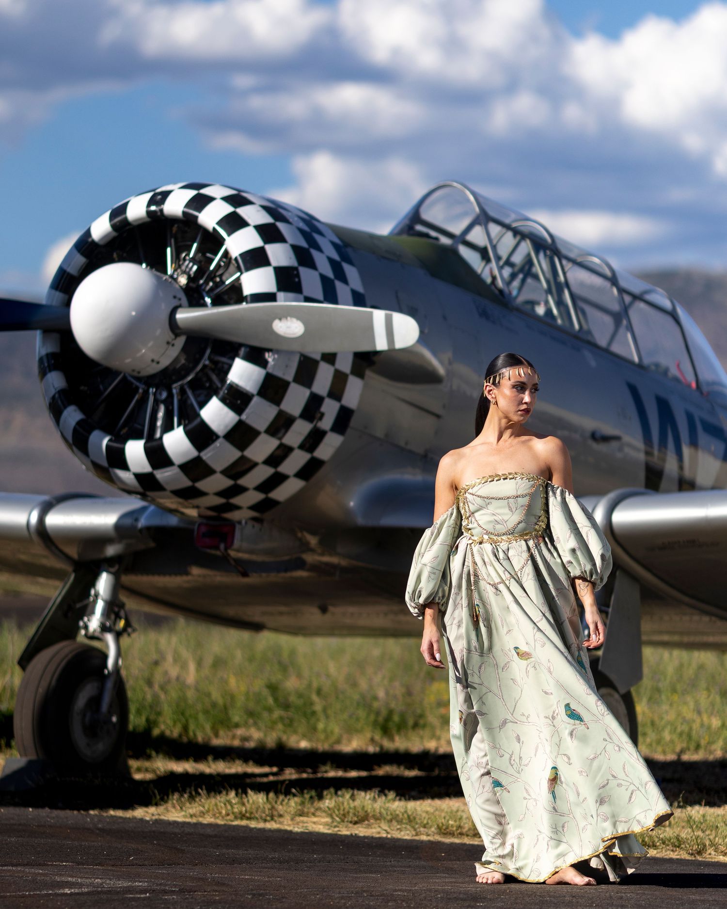 Elegant model in a flowing gown poses beside a vintage checkered propeller aircraft outdoors.