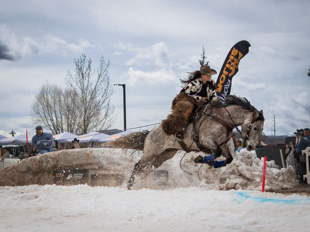 Cowboy riding horse through snow at winter rodeo event with banner and spectators.