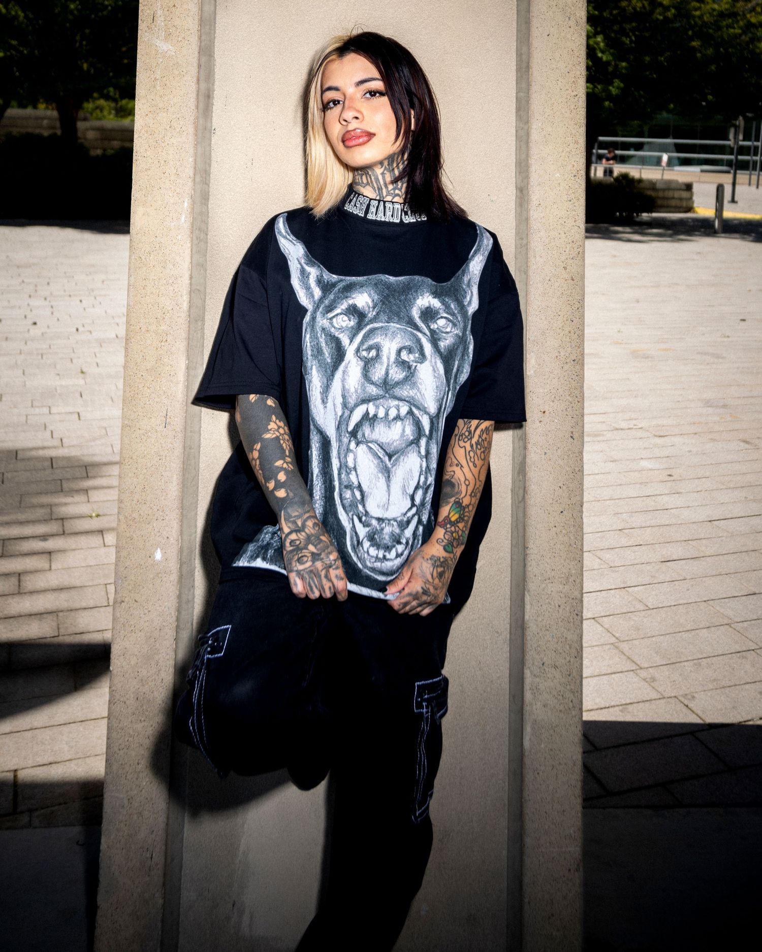 Tattooed woman in oversized black Doberman graphic tee and cargo pants leaning against concrete pillar outdoors.