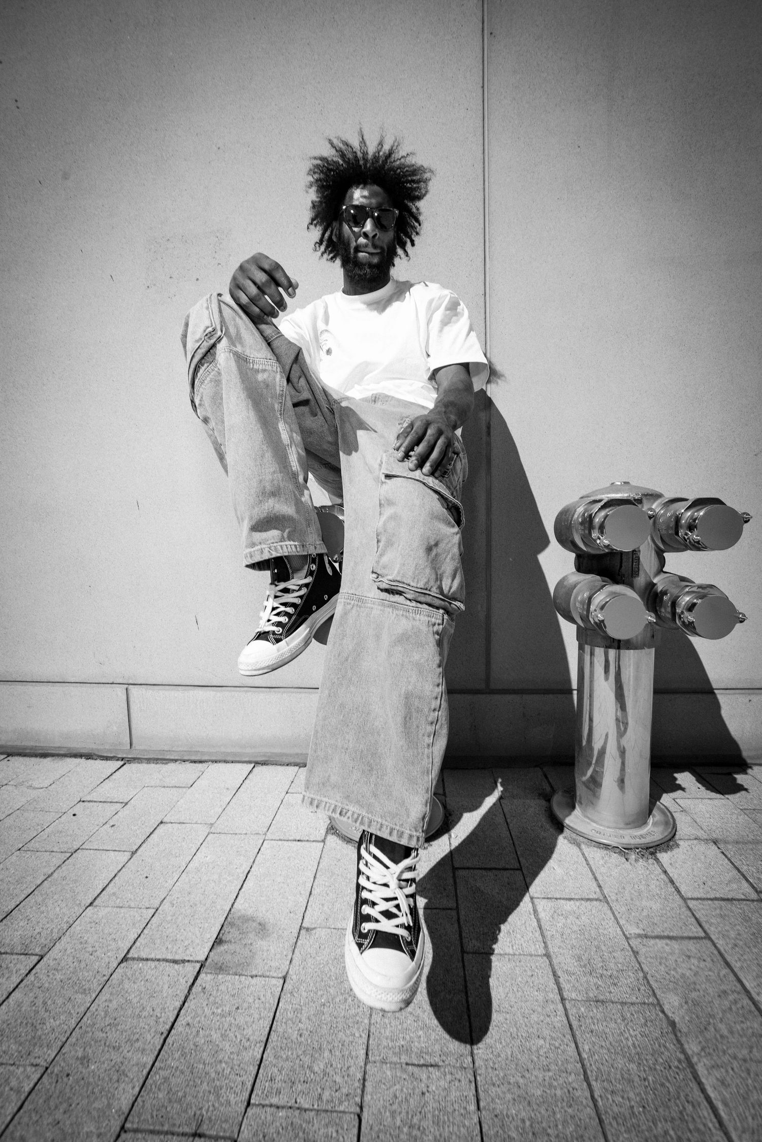 Black and white photo of a man with afro hair, sunglasses, white tee, baggy jeans, and Converse sneakers posing near a fire hydrant.