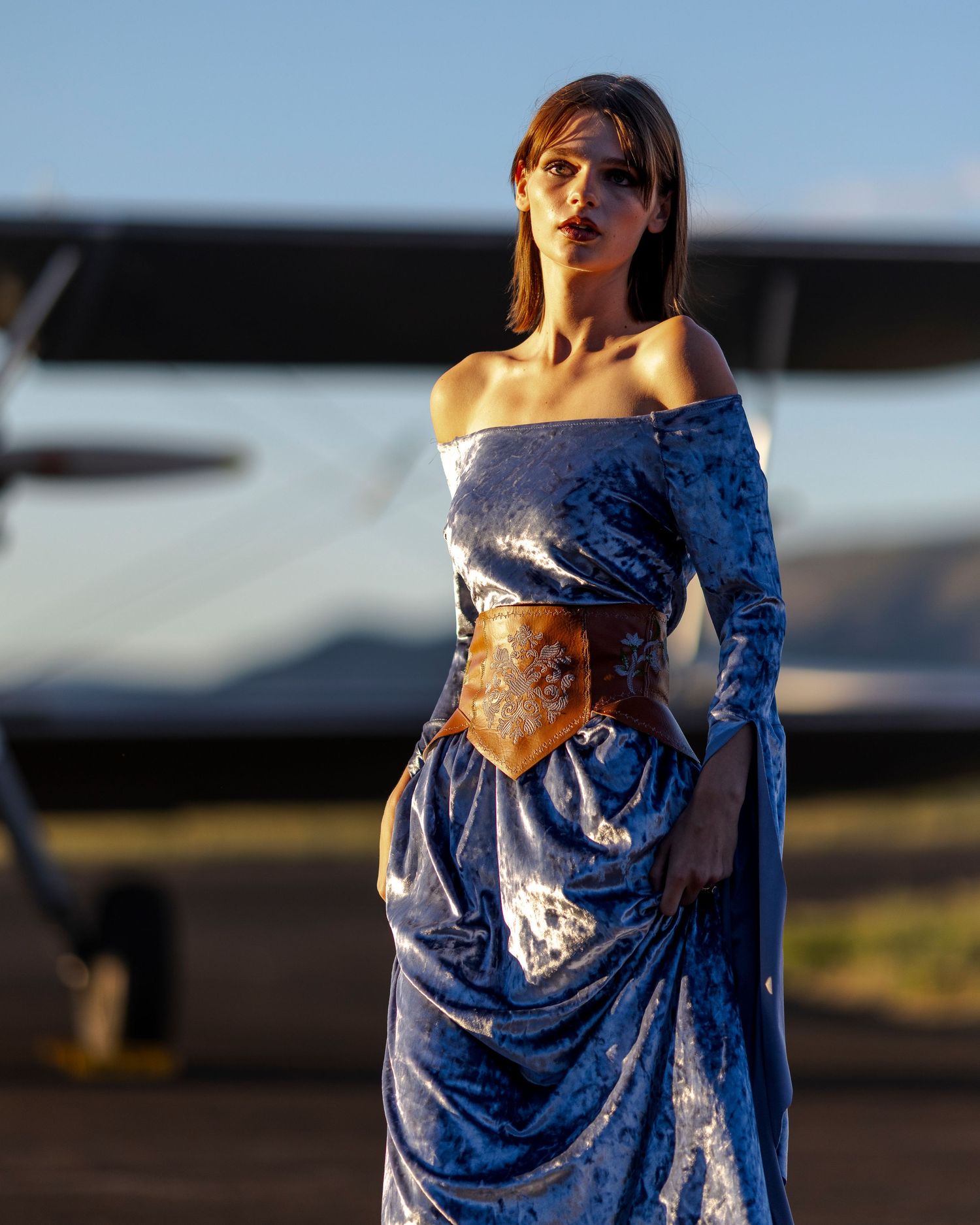Model in blue velvet off-shoulder gown with leather corset belt poses before aircraft at dusk.