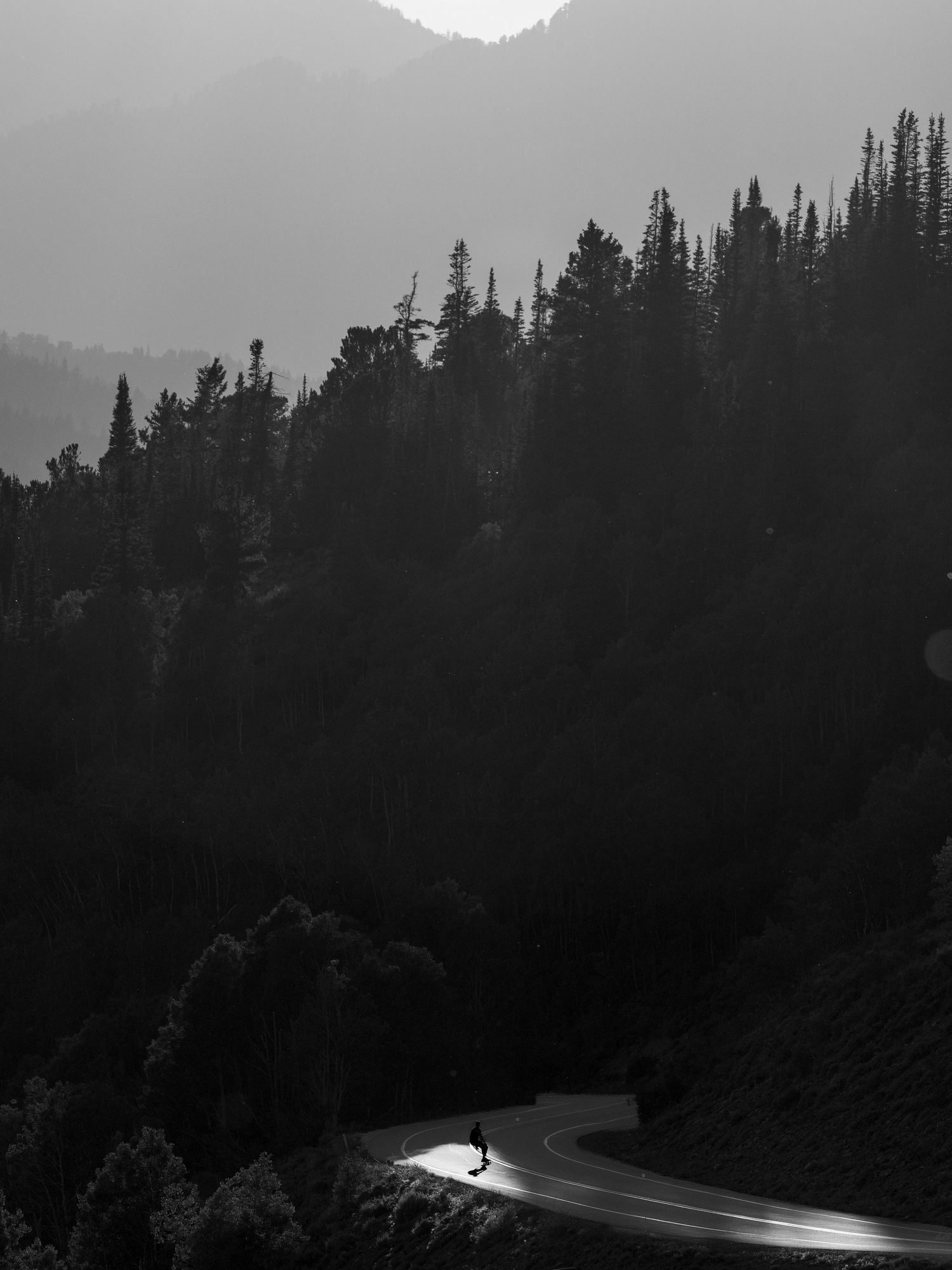 Black and white photo of a lone skateboarder on a winding mountain road at dusk.