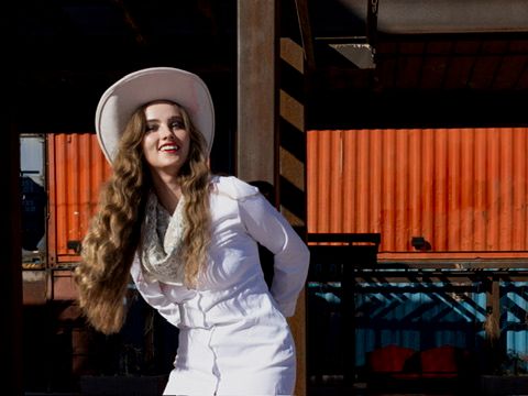 Young woman in white outfit and cowboy hat posing near colorful shipping containers.