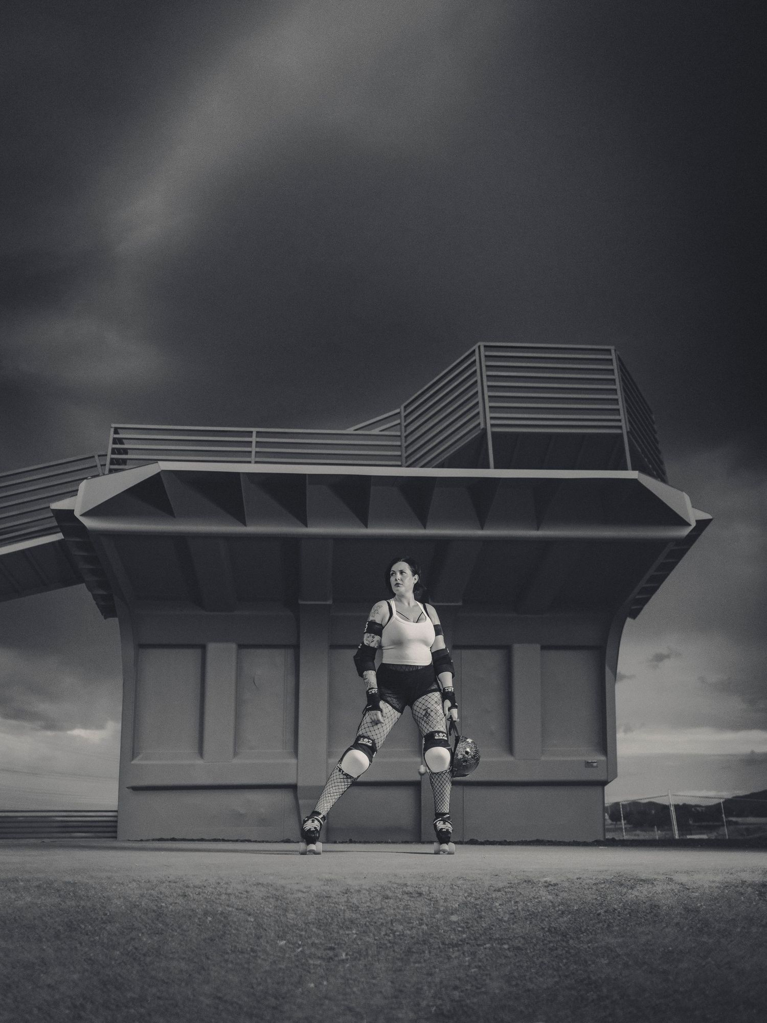 Black and white photo of a roller derby skater posing beneath a structure under stormy skies.