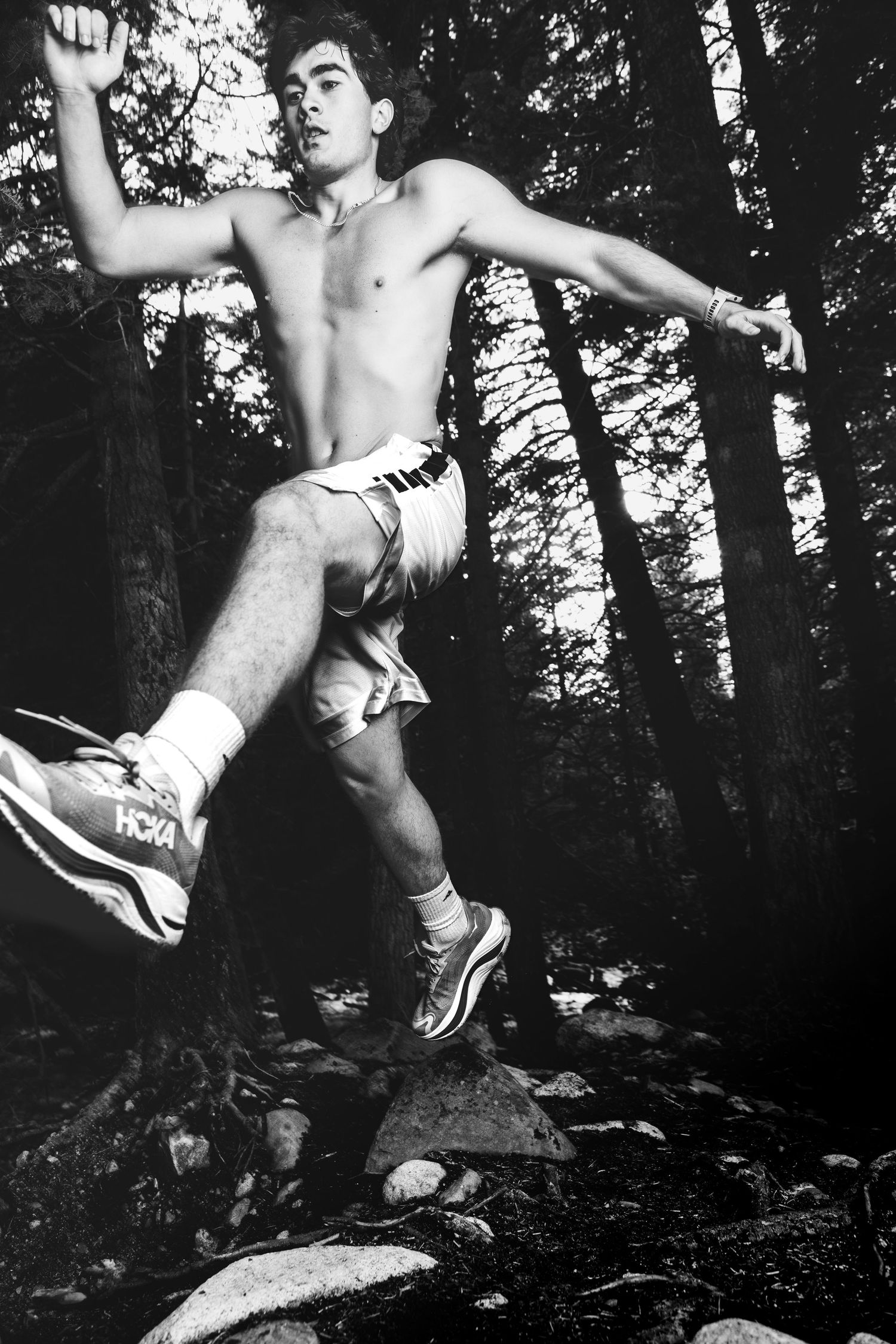 Black and white photo of shirtless male runner leaping over rocks in a forest wearing Hoka sneakers and shorts.