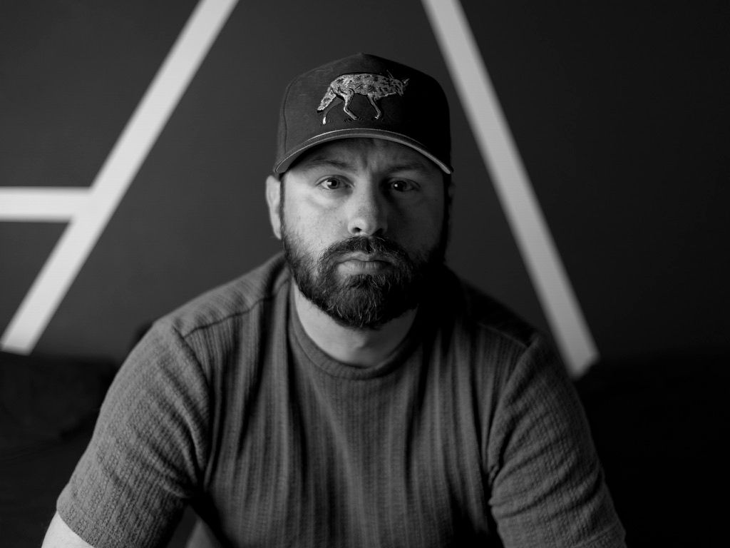 Black and white portrait of bearded man in baseball cap seated against geometric background.