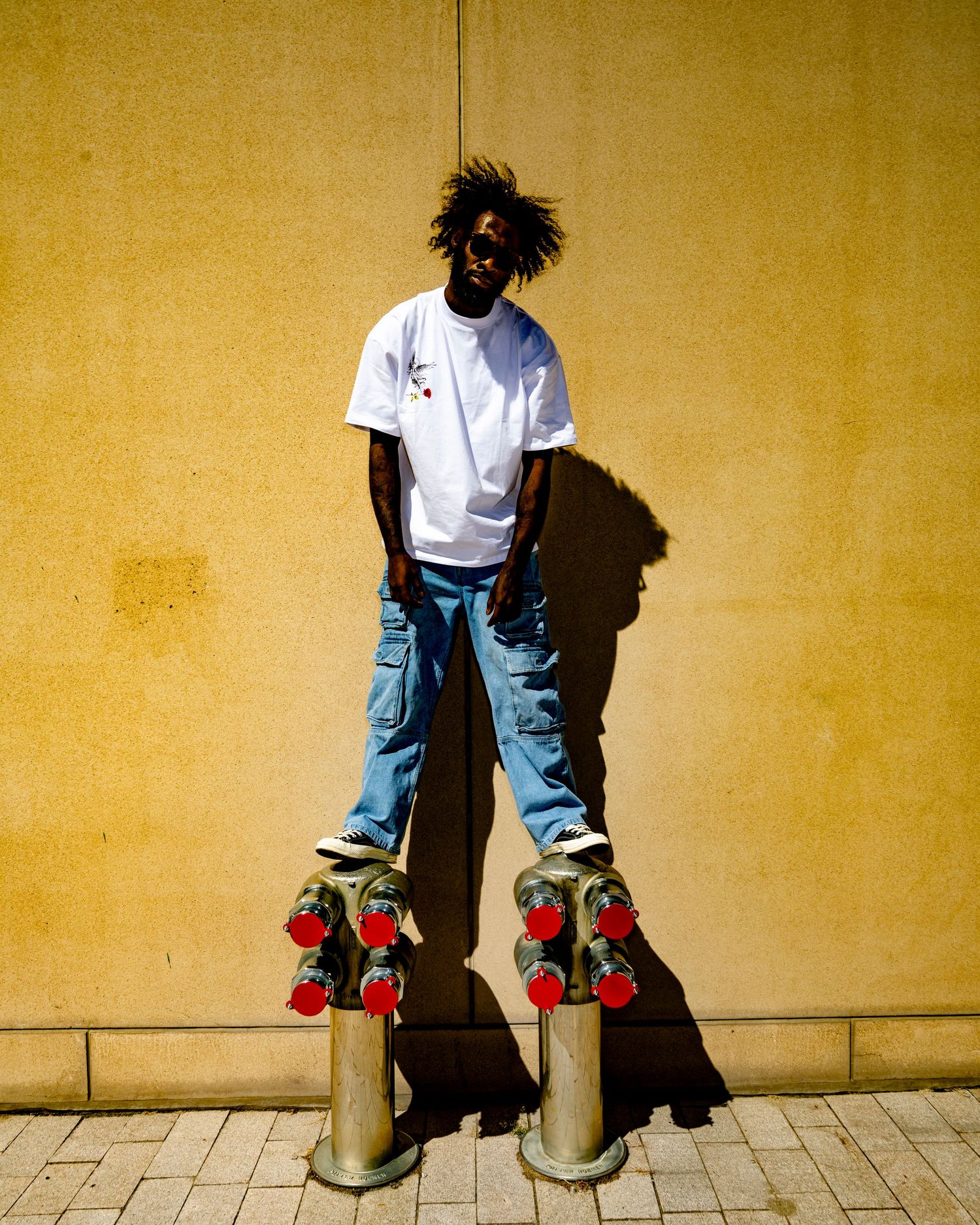 Young man in white tee and blue jeans standing on bollards against a yellow wall outdoors.