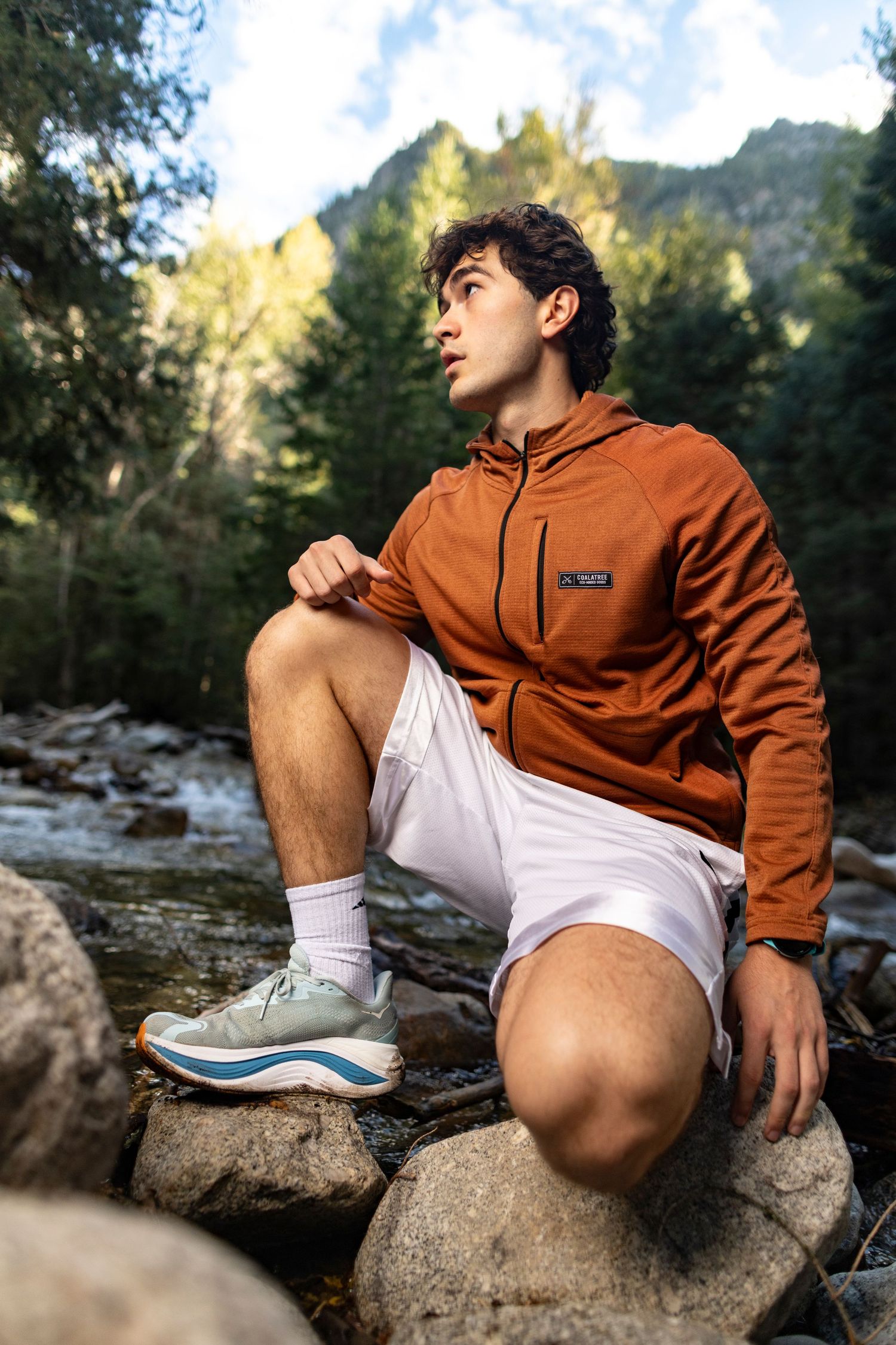 Young man in orange jacket and white shorts sitting on rocks by a mountain stream in a forest setting.