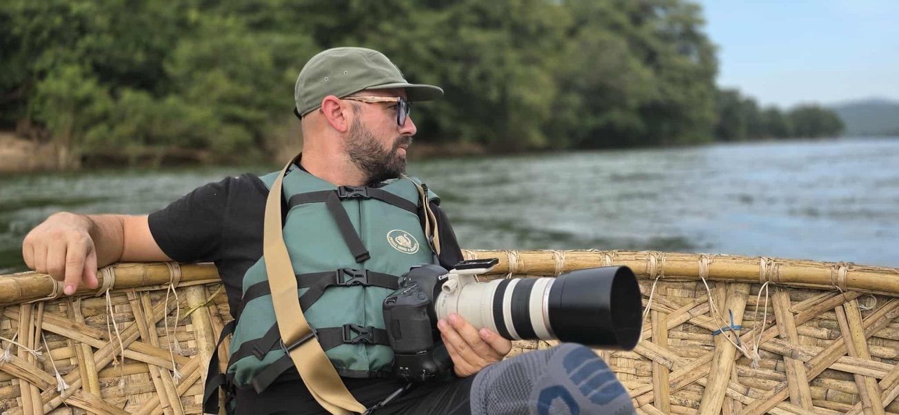 A wildlife photographer with a telephoto lens sits in a bamboo boat on a lush tropical river.