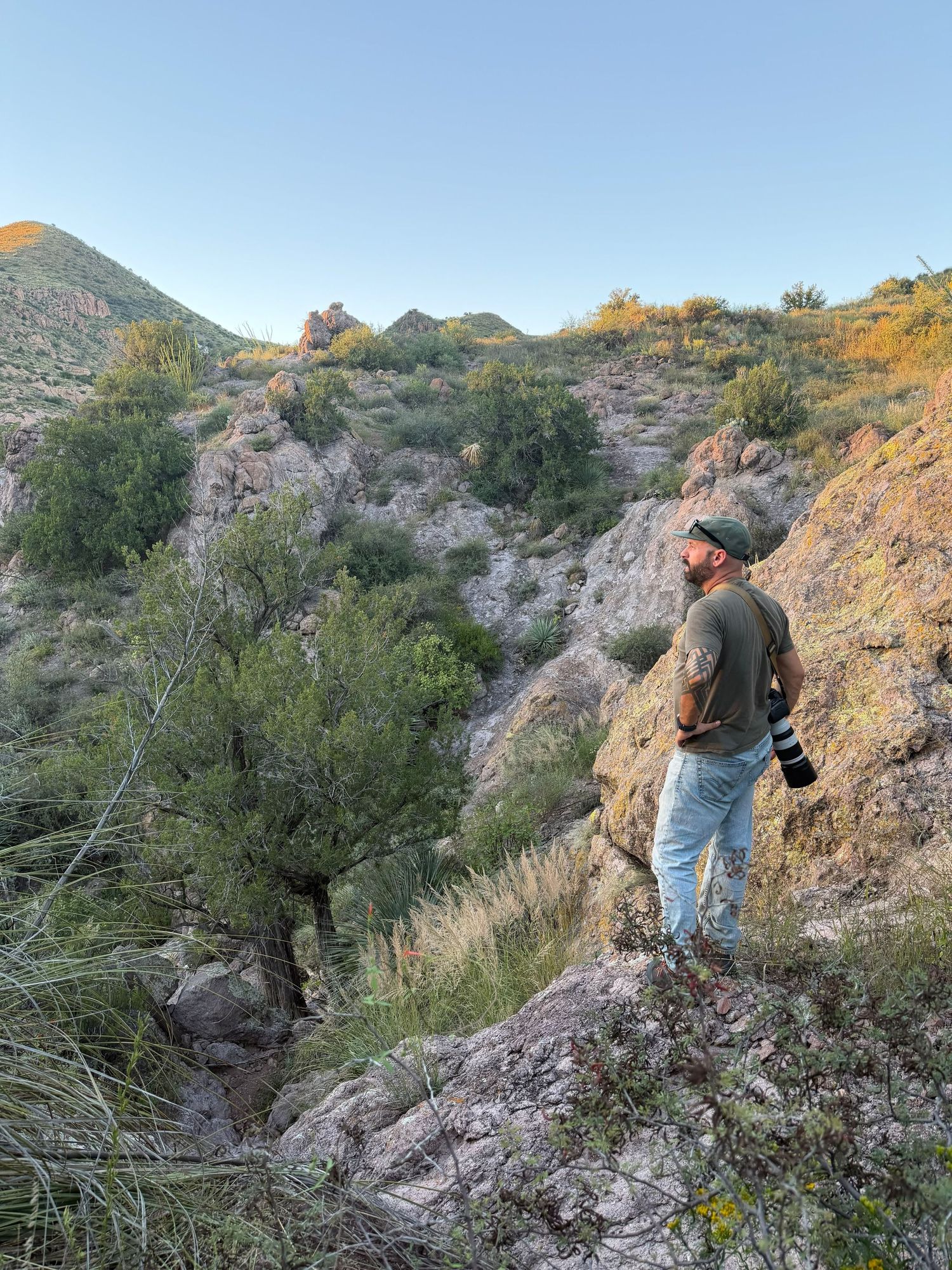 A photographer stands on rocky terrain in a desert canyon, holding a camera while surveying the rugged landscape at dusk.