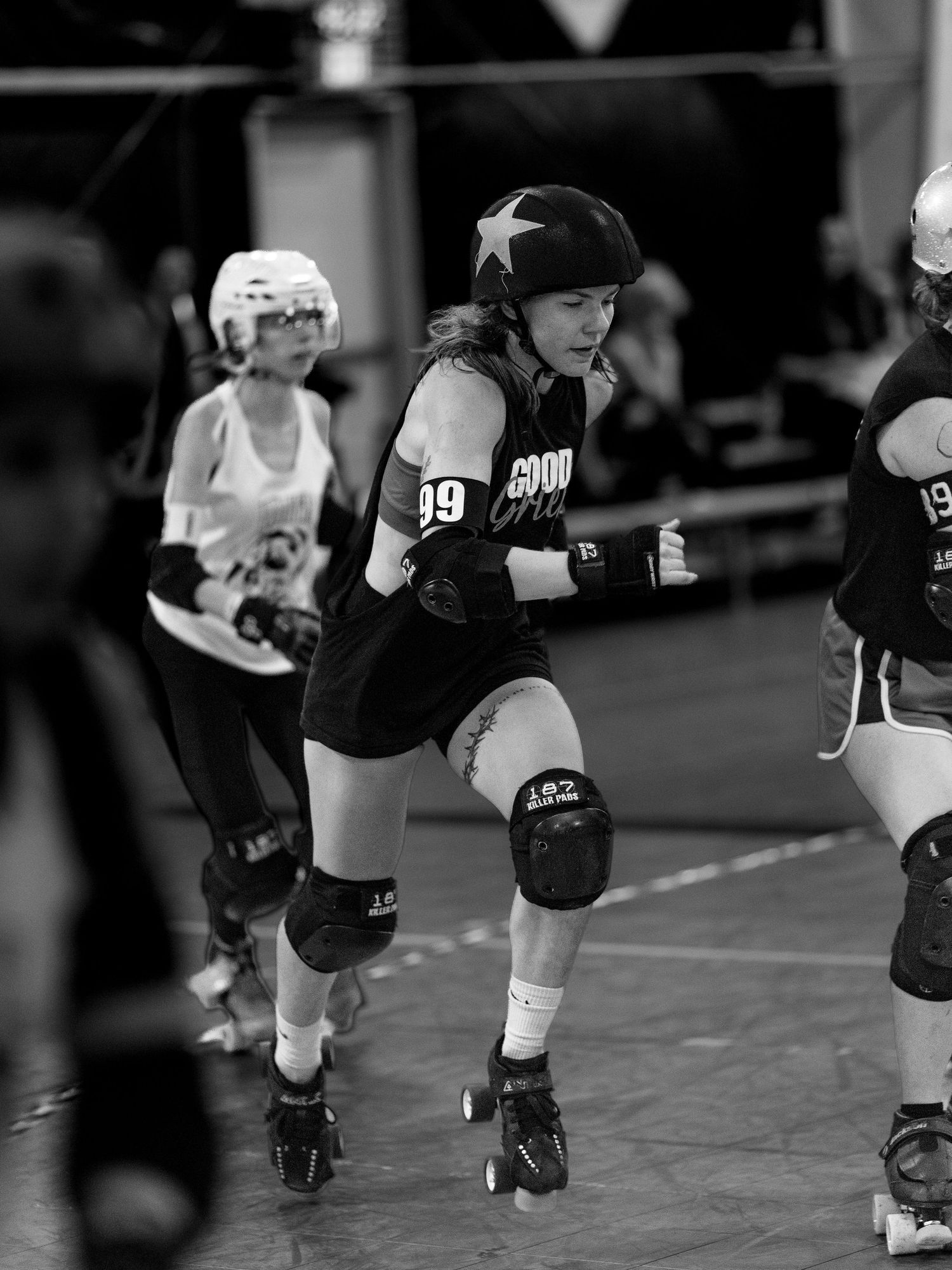 Black and white photo of roller derby players in action, wearing helmets and knee pads, competing on a skating rink.
