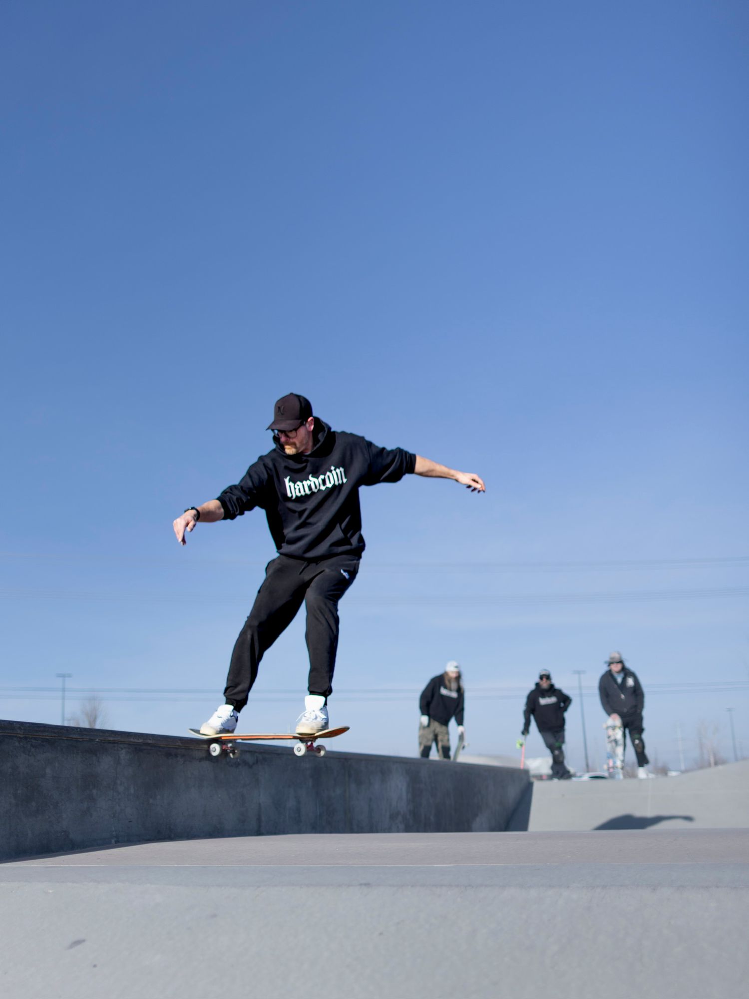 Skateboarder in black hardcoin hoodie performing ollie at skate park under clear blue sky.