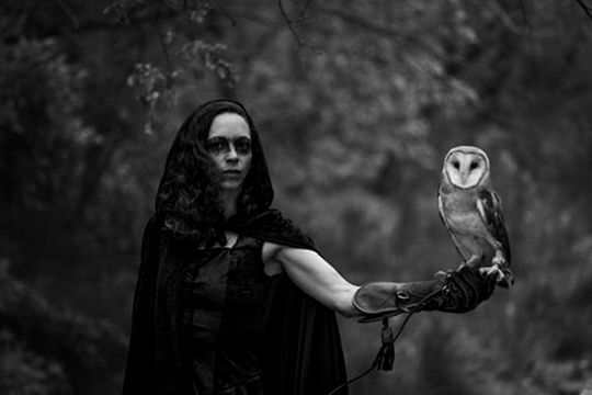 Mysterious woman in black cloak holds barn owl on gloved hand in dark forest black and white photo.