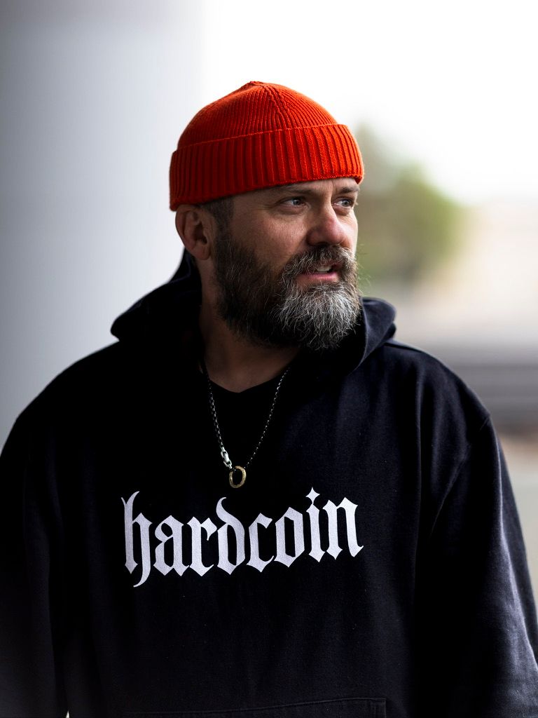 Bearded man wearing red beanie hat and black hardcoin hoodie standing outdoors.
