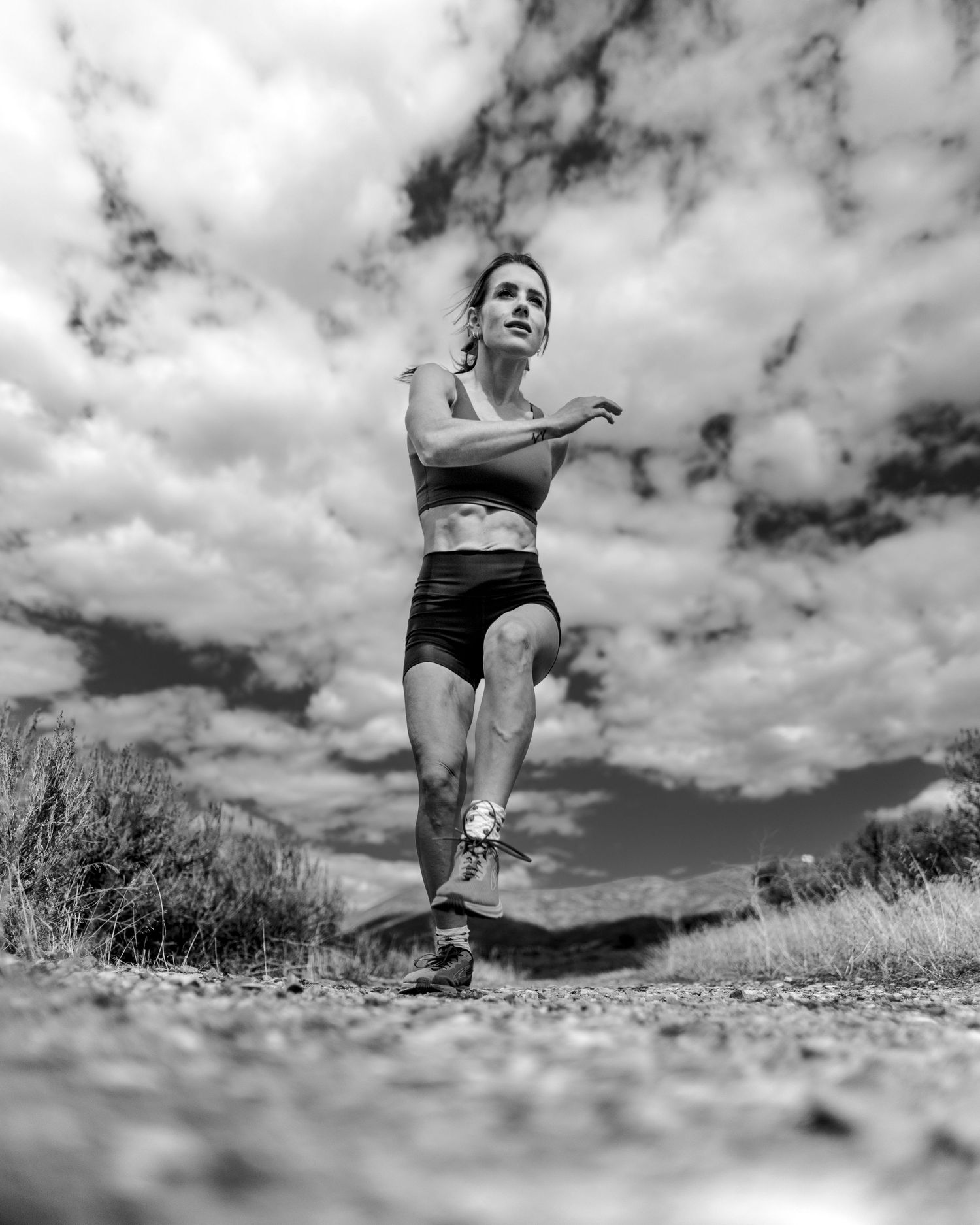 Black and white photo of a fit woman running on a trail path with dramatic cloudy sky in the background.