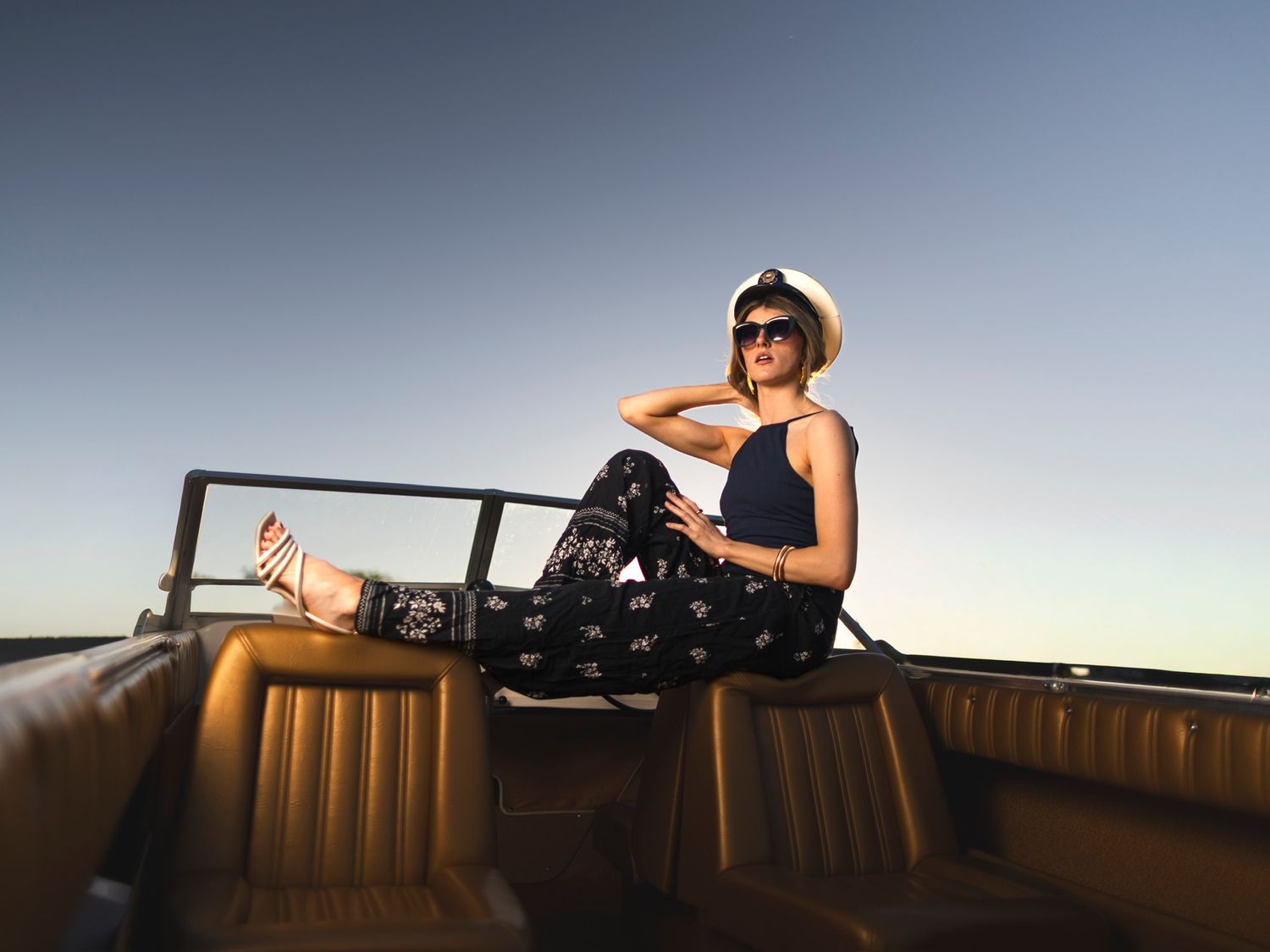 Stylish woman relaxing in vintage convertible car wearing white hat and sunglasses at sunset.