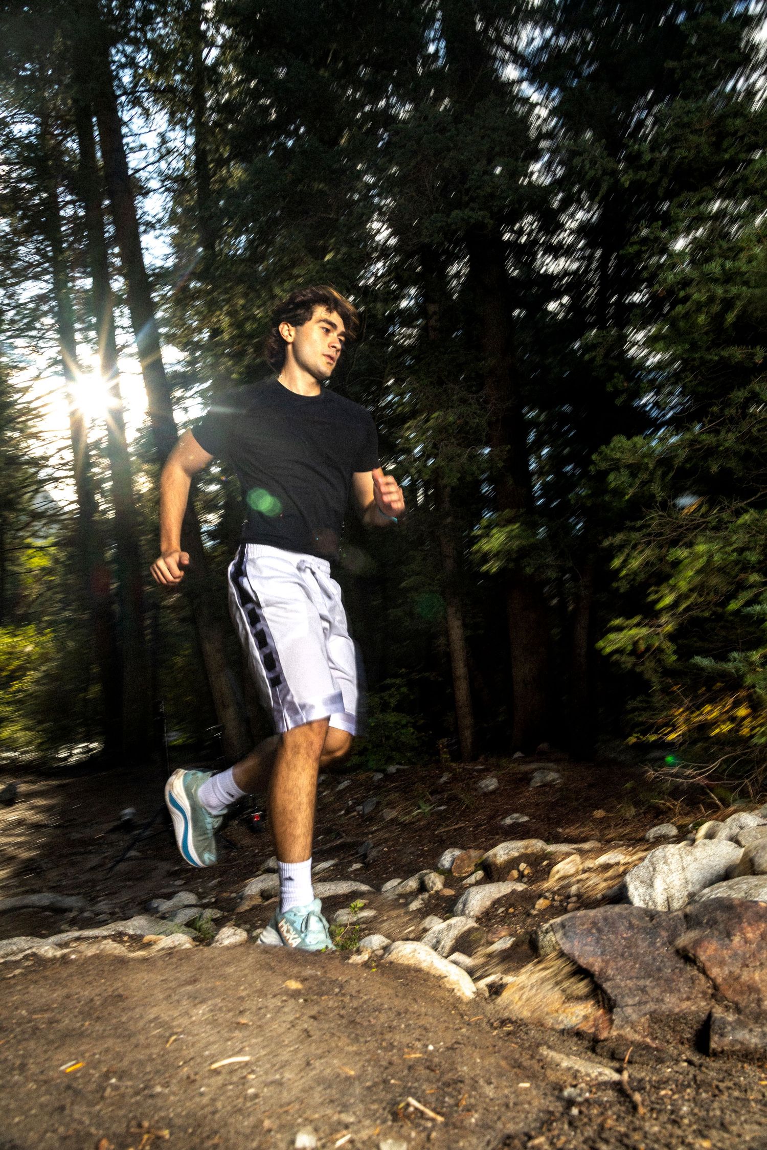 Man in black shirt and white shorts trail running through a sunlit forest on a rocky path.