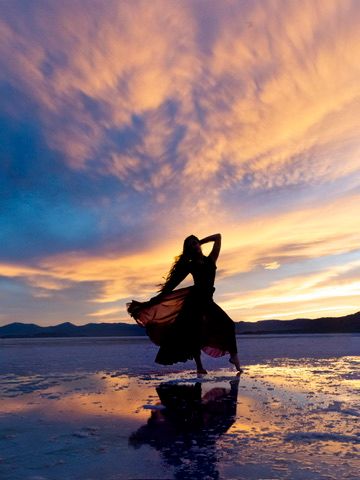 Silhouette of dancer in flowing dress reflected on wet salt flats during vibrant sunset.