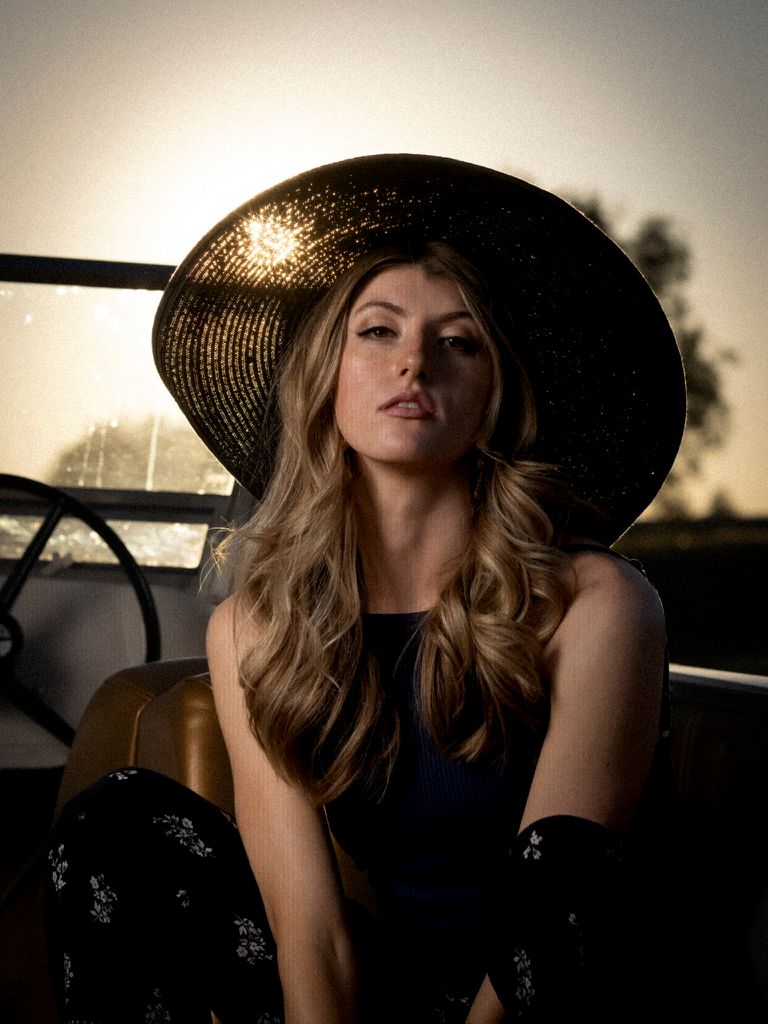 Young woman in a wide-brim black hat sitting outdoors during golden hour sunset light.