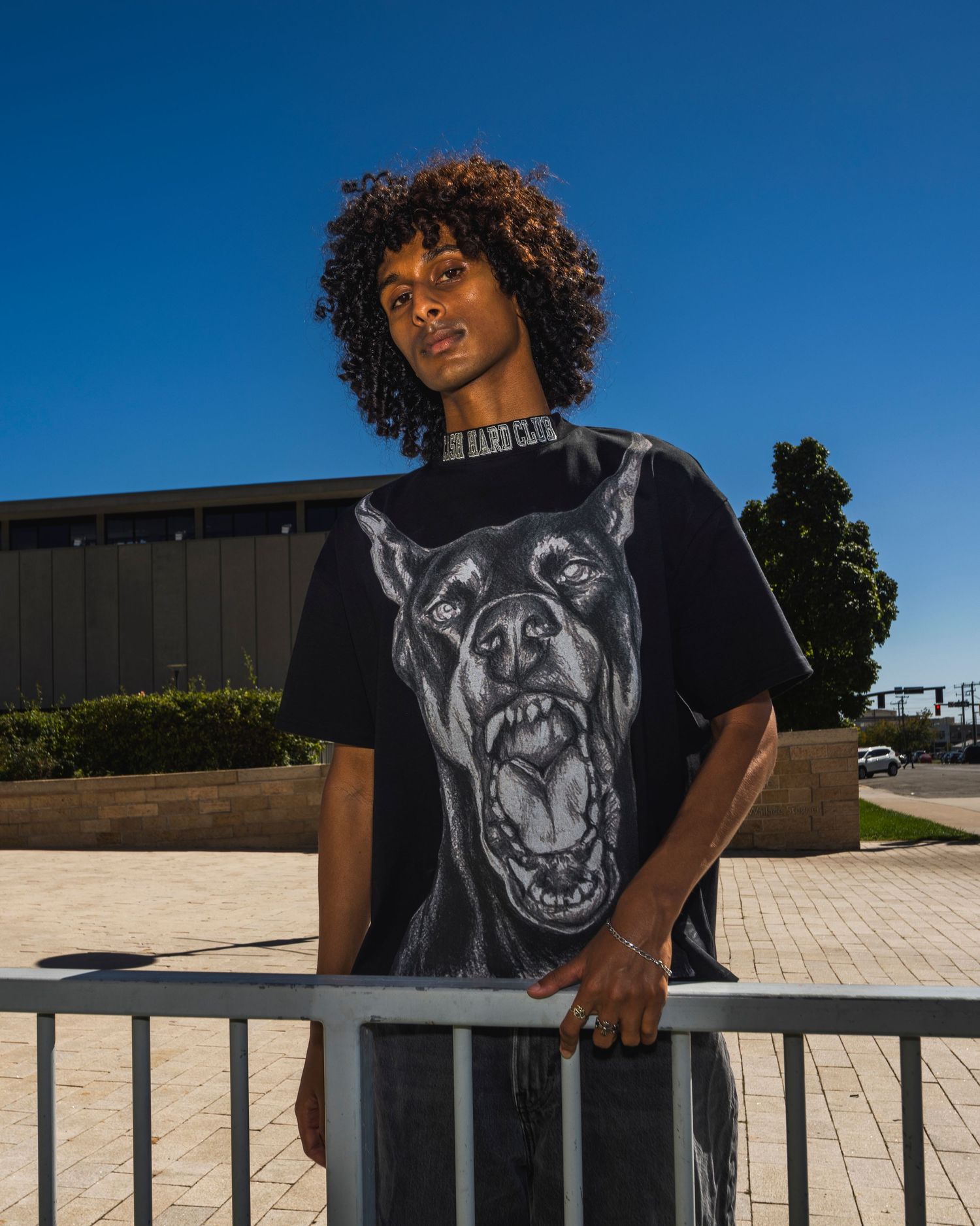 Young man with curly hair wearing an oversized black Doberman graphic t-shirt, leaning on a railing outdoors.