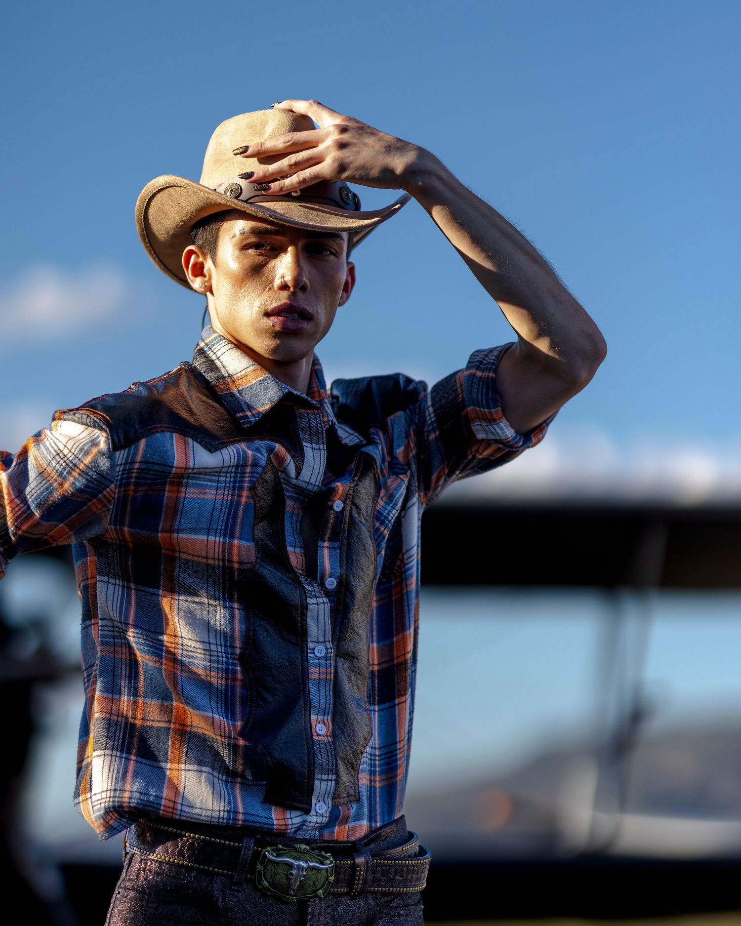 Young man in plaid shirt adjusts cowboy hat against blue sky with aircraft in background.