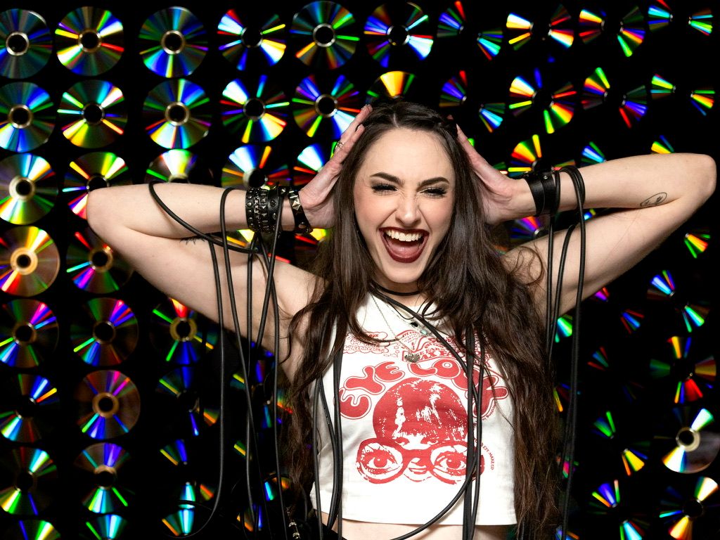 Excited woman in graphic tee poses against colorful CD backdrop with hands in hair.