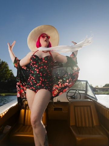 Woman in floral swimsuit and sun hat posing on a wooden boat on a sunny day.
