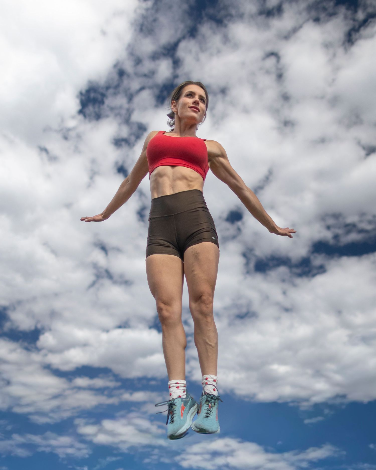 Athletic woman in red sports bra and brown shorts jumps against a blue cloudy sky, shot from below.