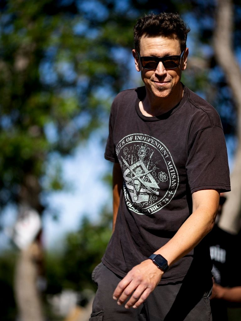 Smiling man in sunglasses and graphic t-shirt walking outdoors on a sunny day.