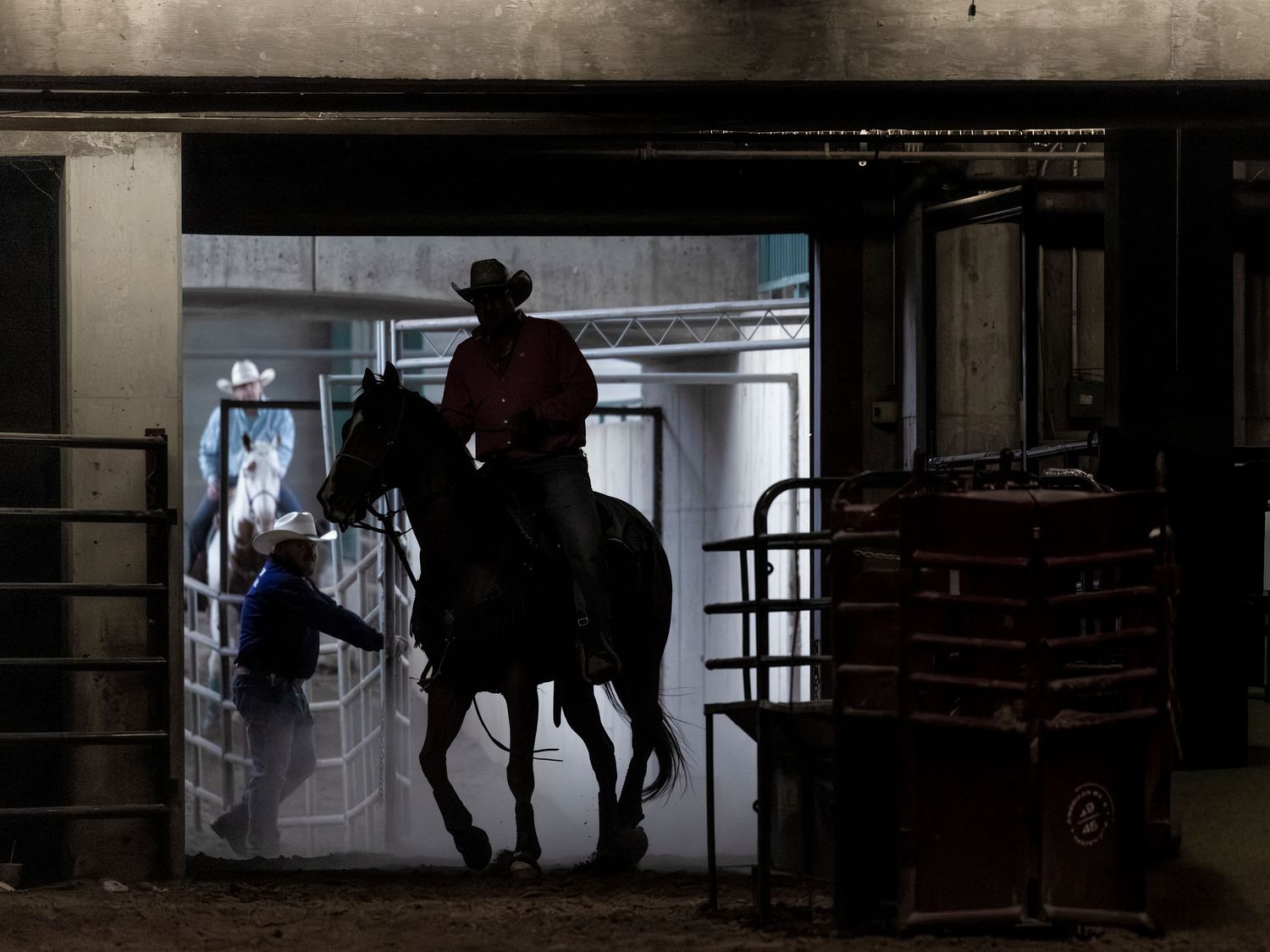 Silhouette of a cowboy on horseback emerging from a dark arena tunnel into dim light.