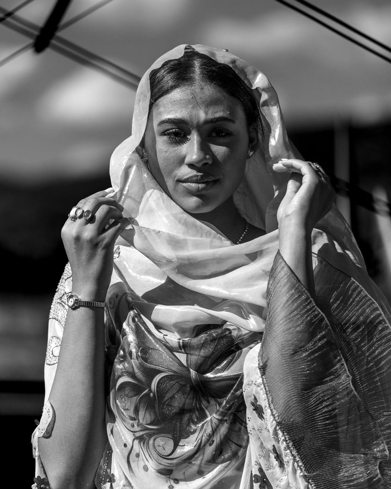 Black and white portrait of model wearing sheer head scarf and patterned dress in natural light.