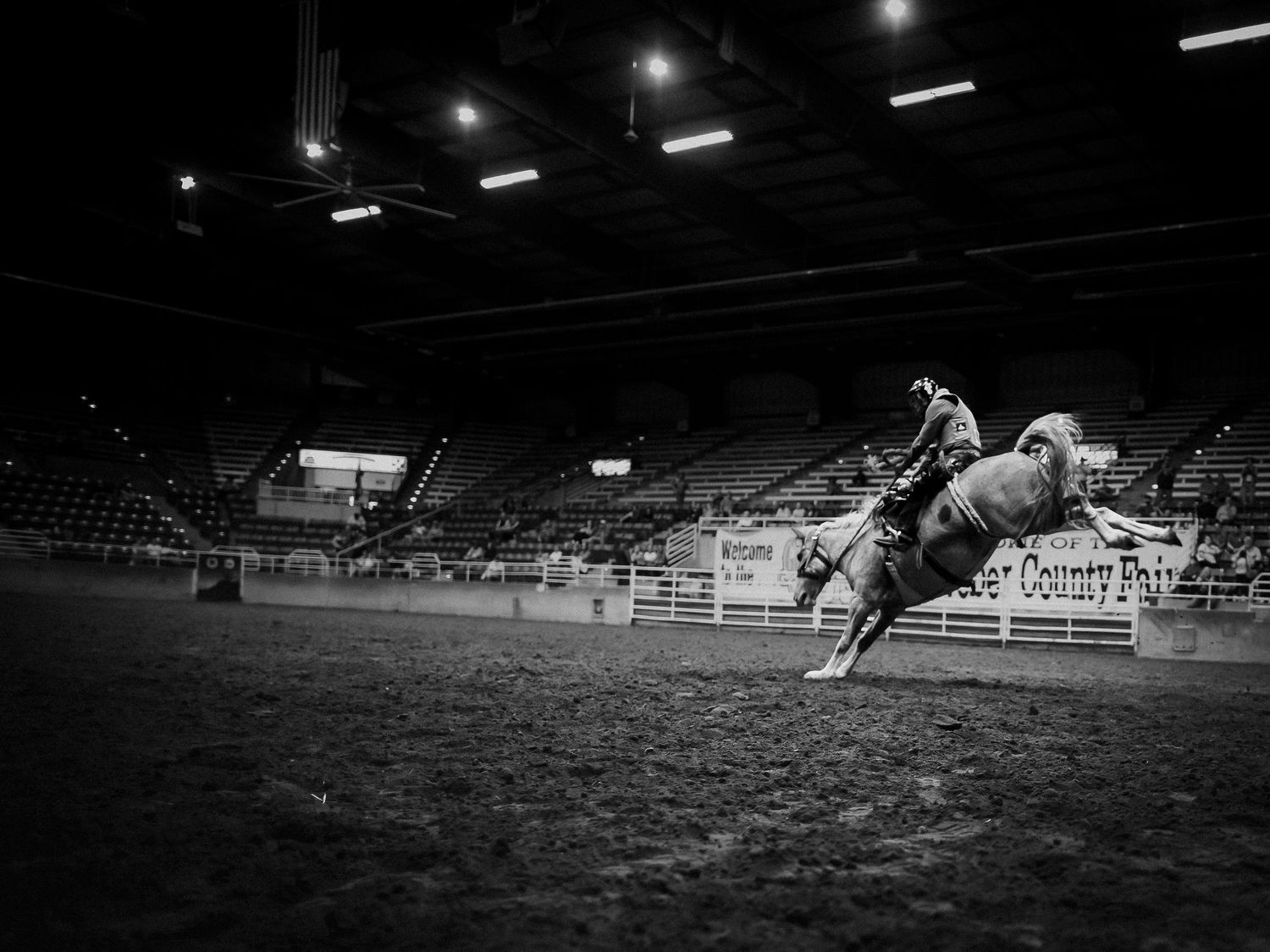 Black and white photo of a rodeo rider on a bucking horse in an indoor arena.