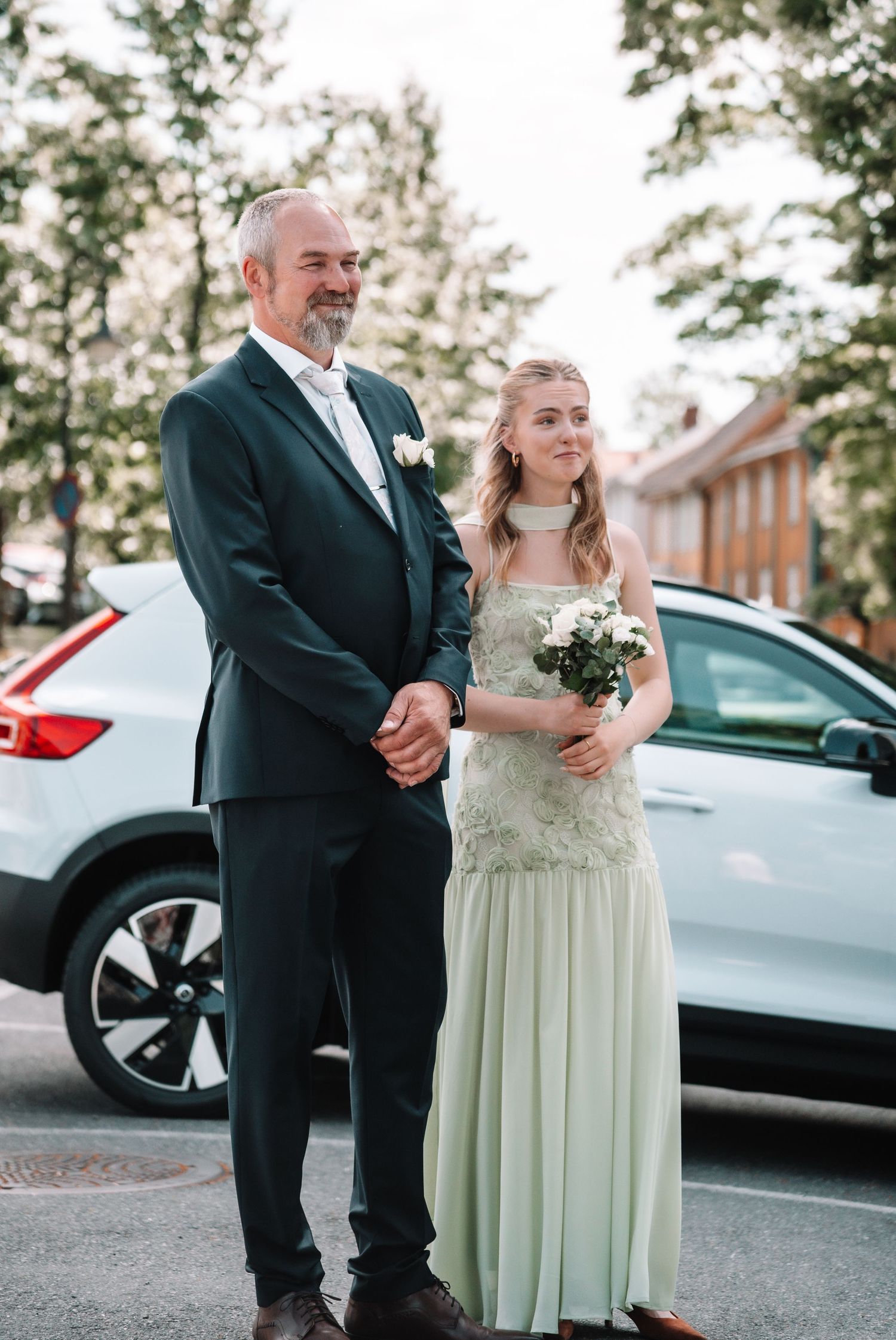Father in dark suit proudly standing beside young bridesmaid in sage green dress holding white bouquet.