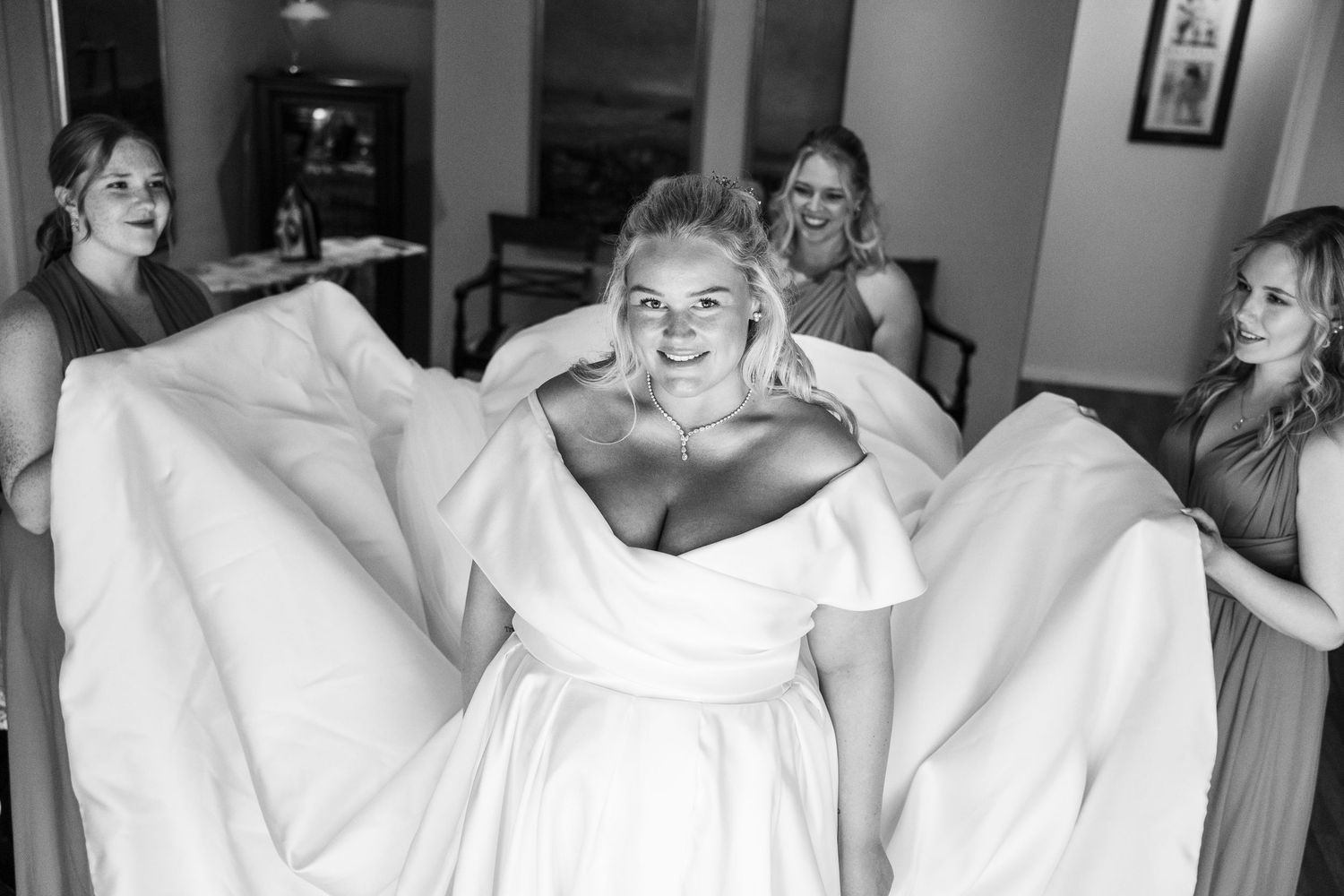 Black and white photo of bridesmaids spreading train of brides white wedding gown while she smiles joyfully.