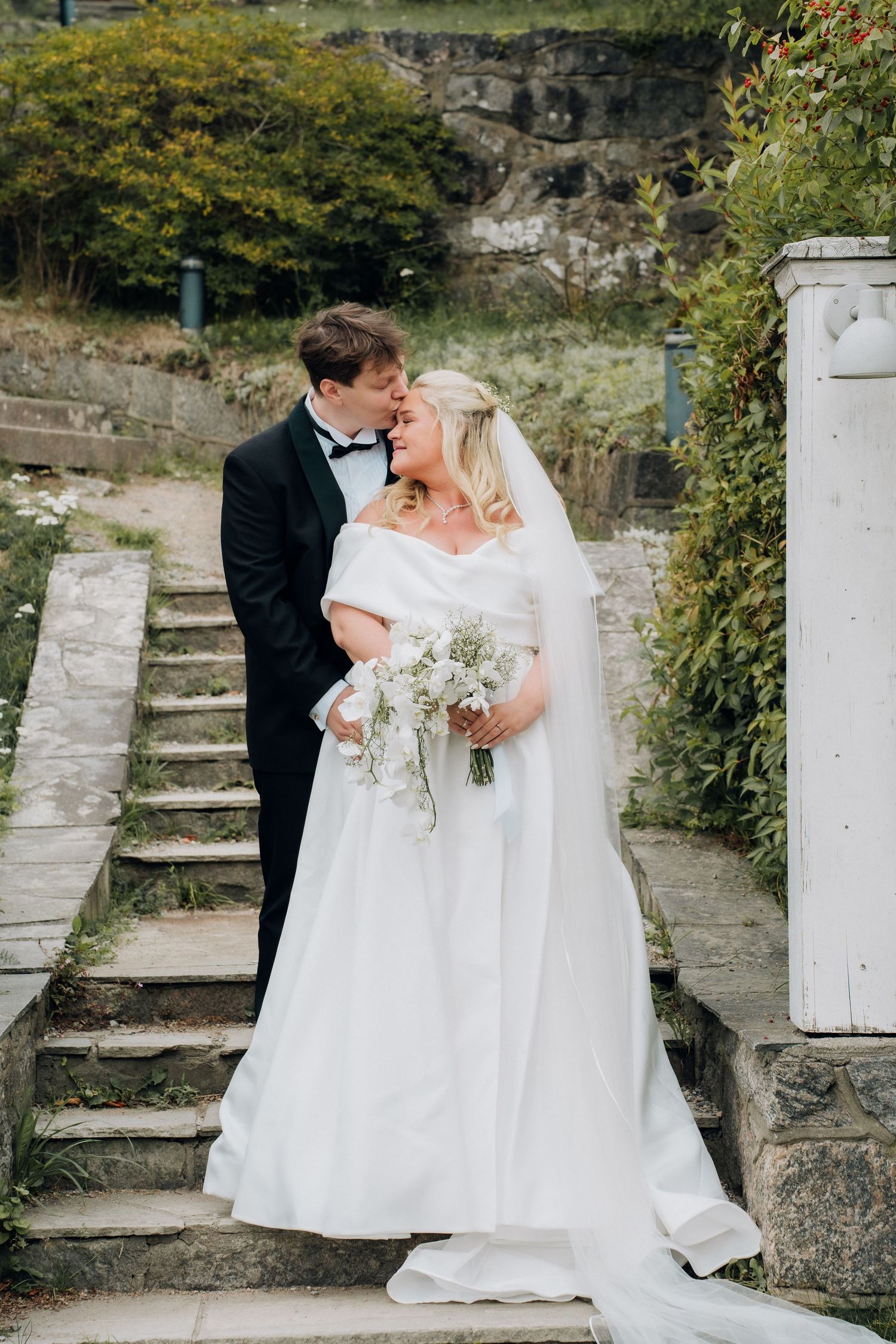 Bride and groom sharing a kiss on stone steps surrounded by greenery, bride holding white bouquet.