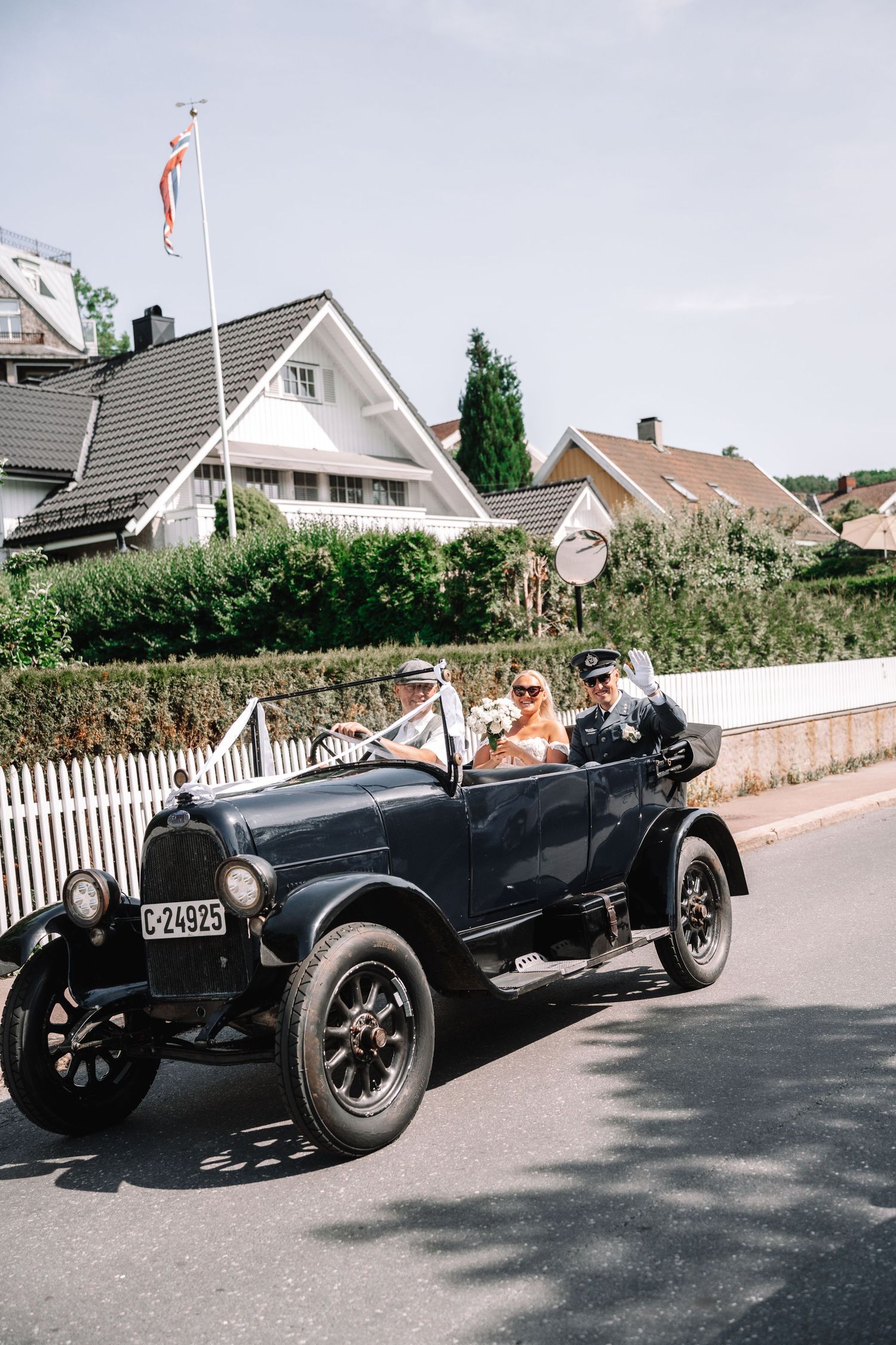 Bride and groom riding in a vintage black convertible car through a residential street on their wedding day.