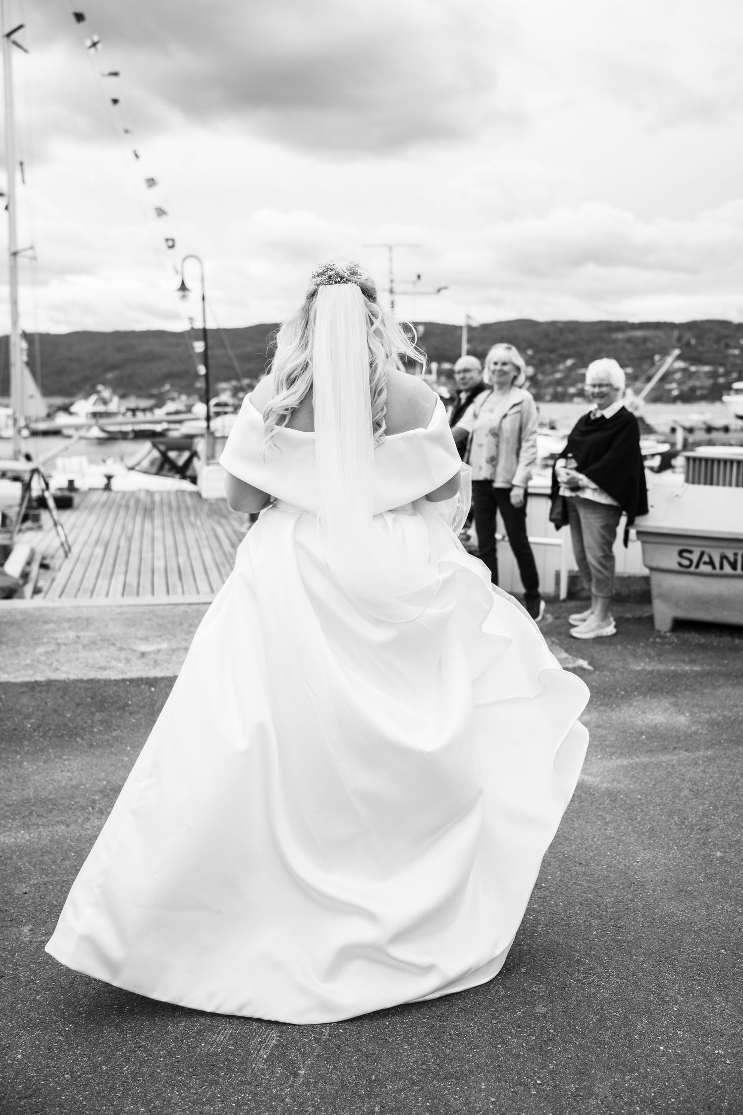 Bride in white gown and veil walking toward guests at a waterfront marina.