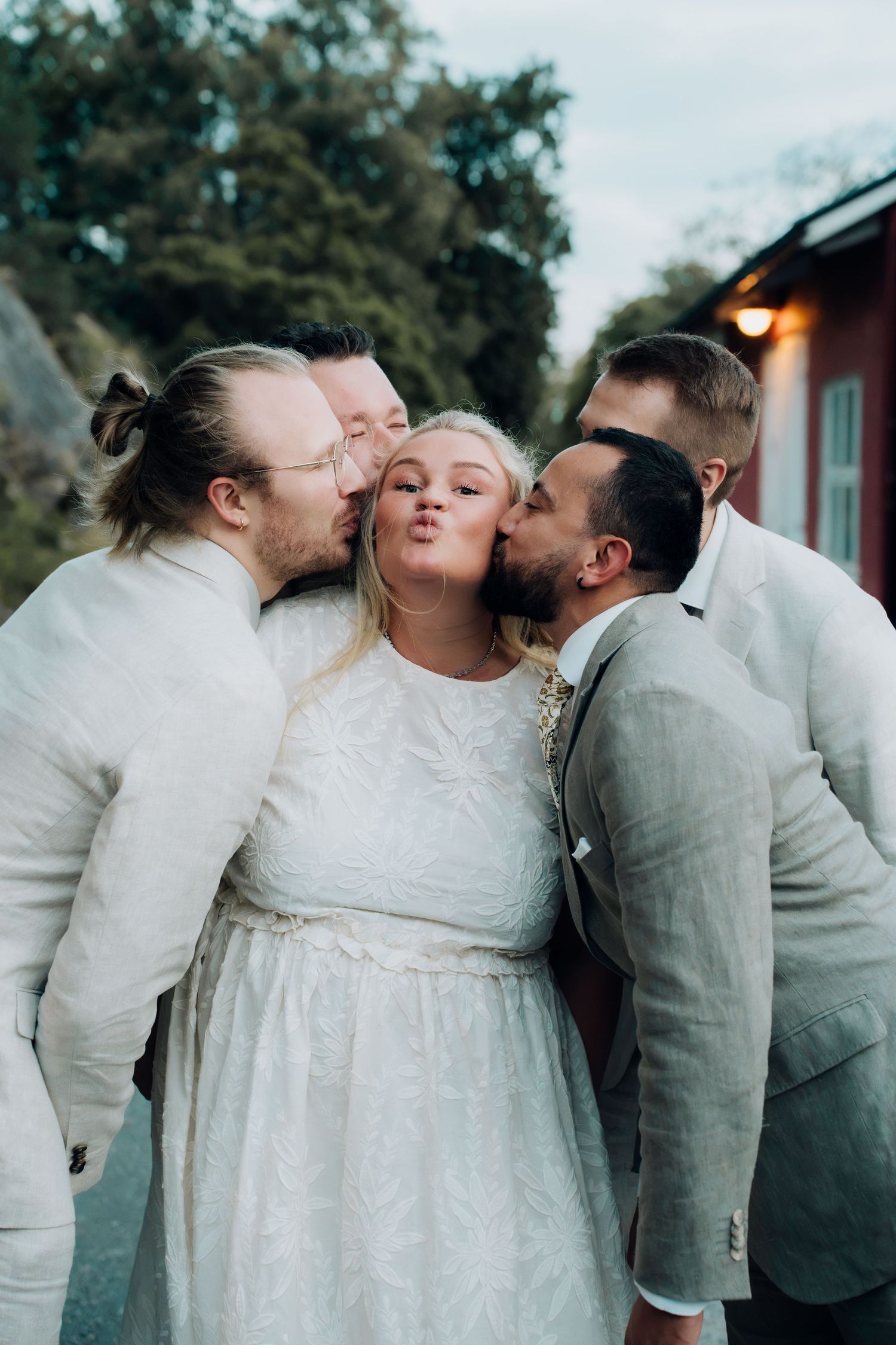 Bride in white dress receives kisses on cheeks from groomsmen in linen suits at outdoor wedding.