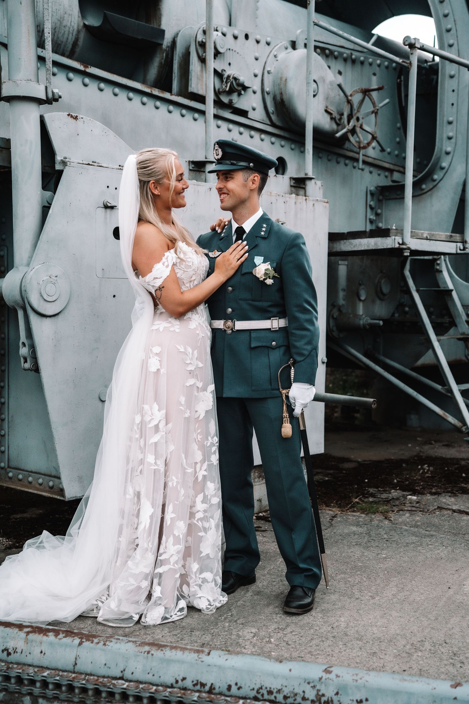 Brud i blomstrete blondekjole og brudgom i militæruniform poserer sammen foran et veterantog.