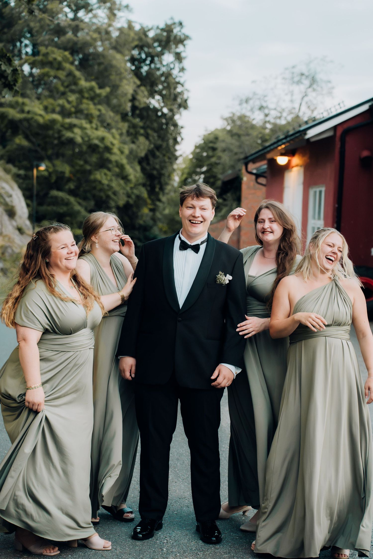 Groom in black tuxedo walks cheerfully with four bridesmaids in sage green gowns outdoors.