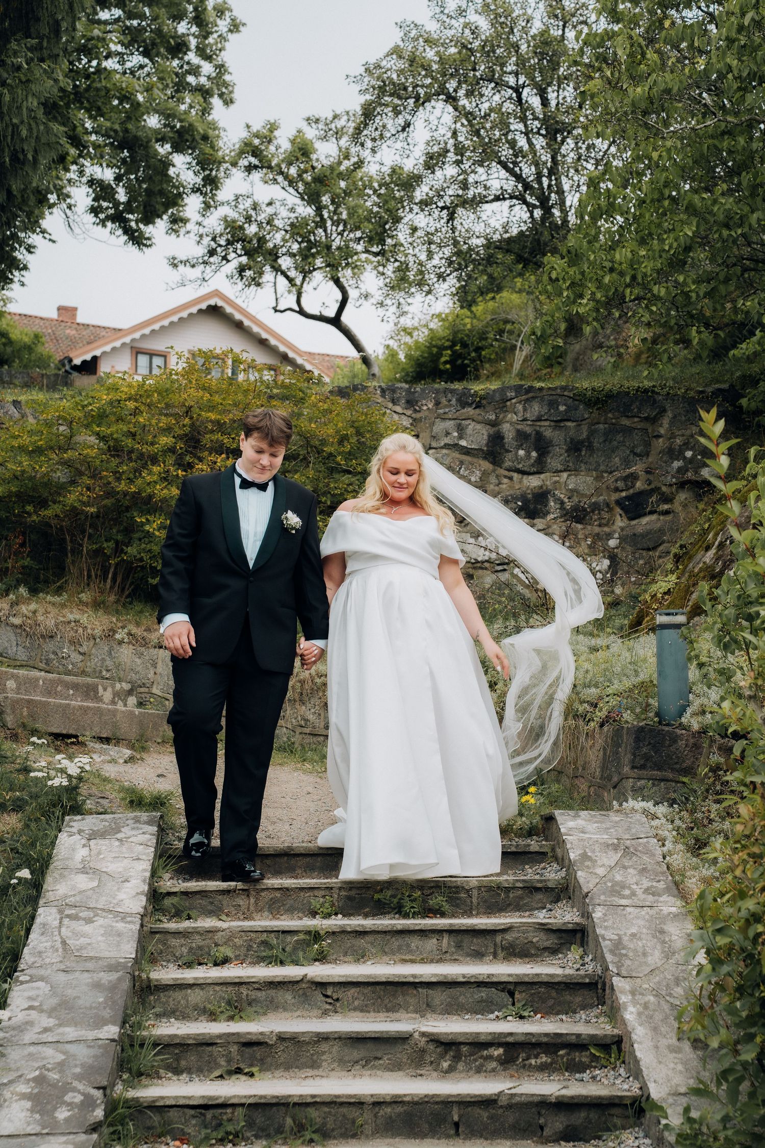 Bride and groom holding hands descending stone steps in a lush garden, veil flowing in the wind.