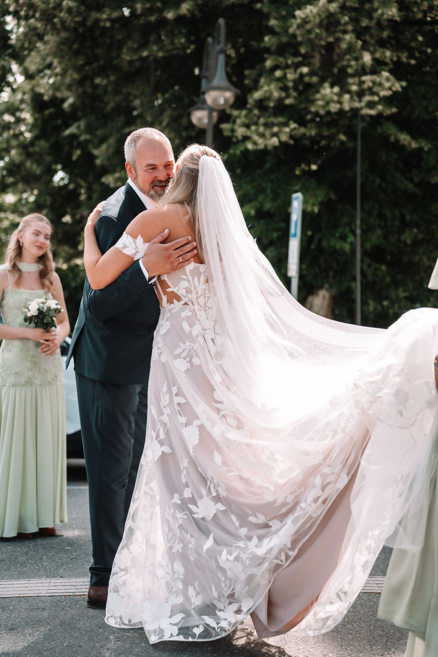 Emotional father embracing his bride daughter in butterfly applique veil outside church with bridesmaids nearby.